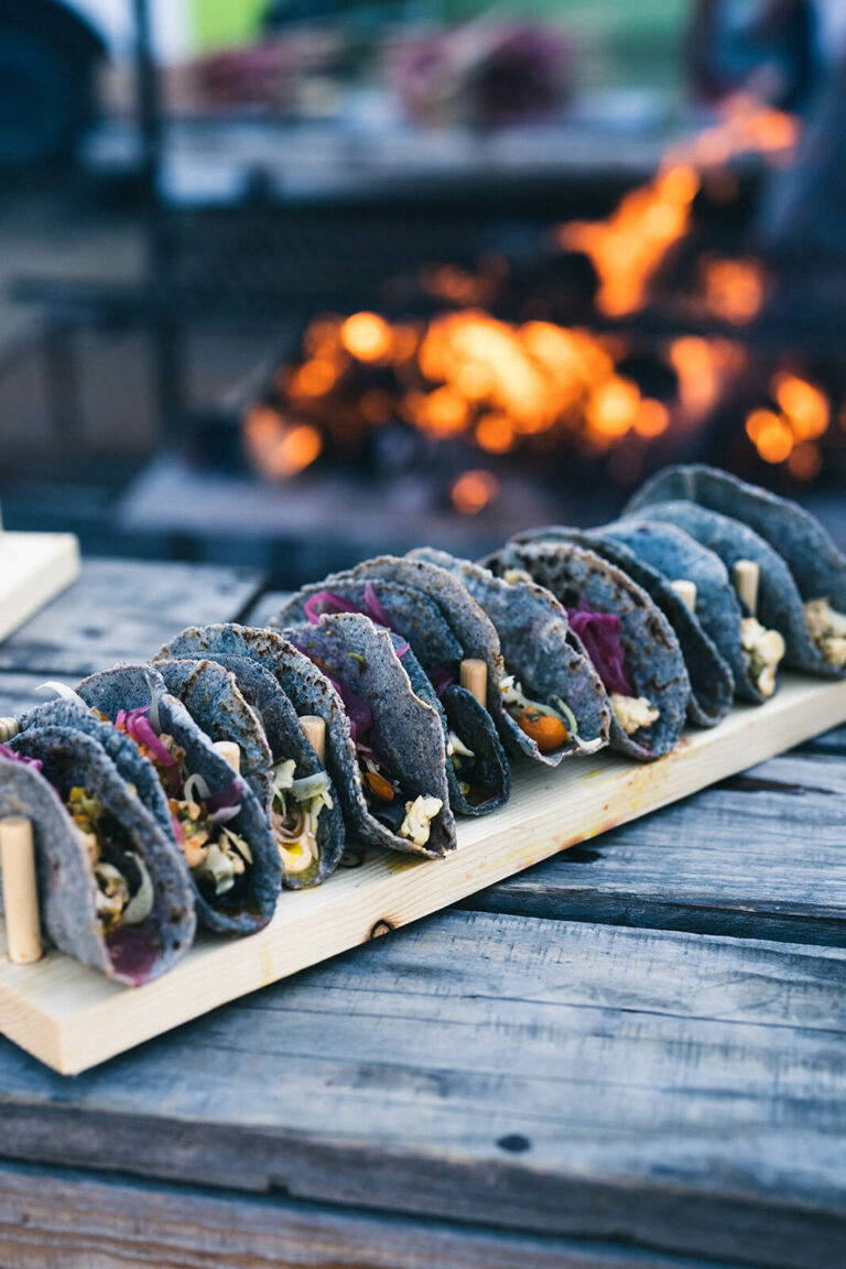 Tacos with blue corn tortillas sit on a table in front of an open fire.