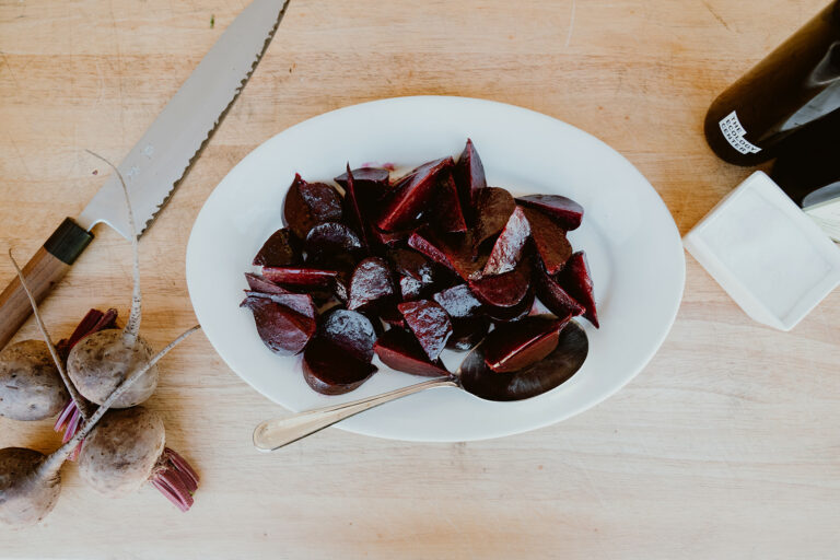 Marinated roasted beets on a plate next to raw beets, knife, and olive oil.