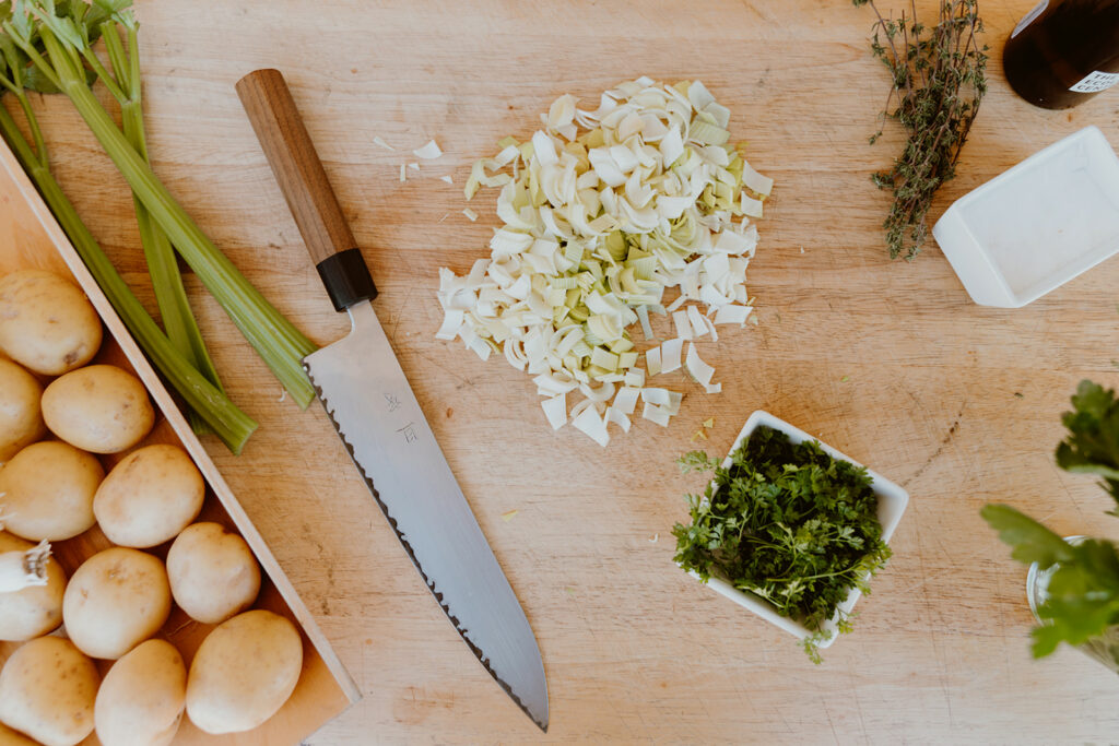 Potatoes, celery, green garlic, parsley, salt and a knife arranged on a cutting board.