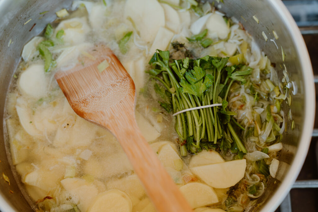 Potatoes, leeks, and green garlic submerged in water with the herb bundles resting on top in a deep pot.
