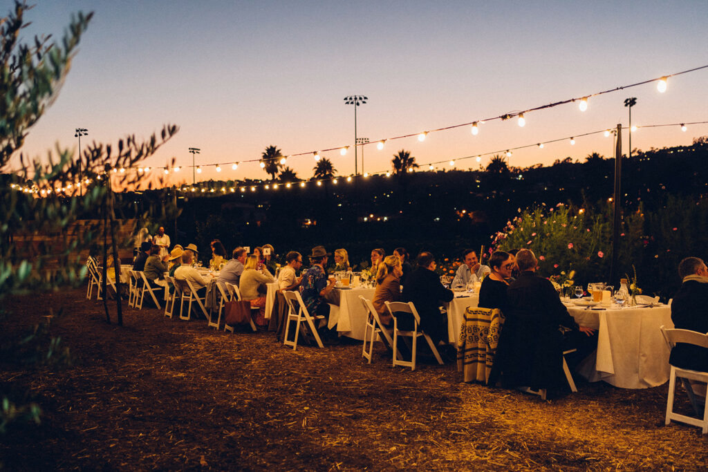 Long row of tables filled with dinner guests and friends in middle of farm field.