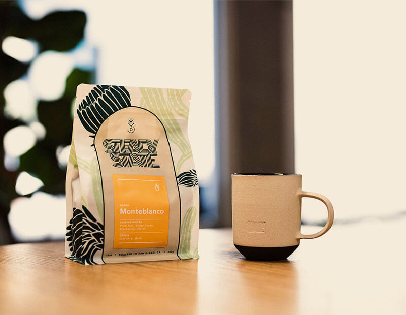 A bag of steady state coffee sits next to a coffee mug on wooden table.